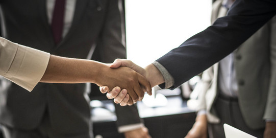 Handschlag Zwei Personen geben sich die Hand. Das Händeschütteln ist im Detail abgebildet, im Hintergrund stehen zwei Menschen im Anzug.