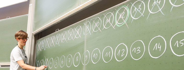 KinderUni-Vorlesung Dozentin und Junge stehen vor einer mit Zahlen beschrifteten Tafel im Hörsaal