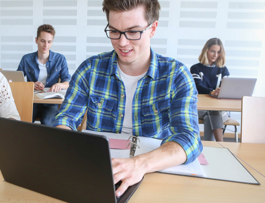 Studierender mit Laptop Student sitzt im Seminarraum am Laptop, im Hintergrund weitere Studierende